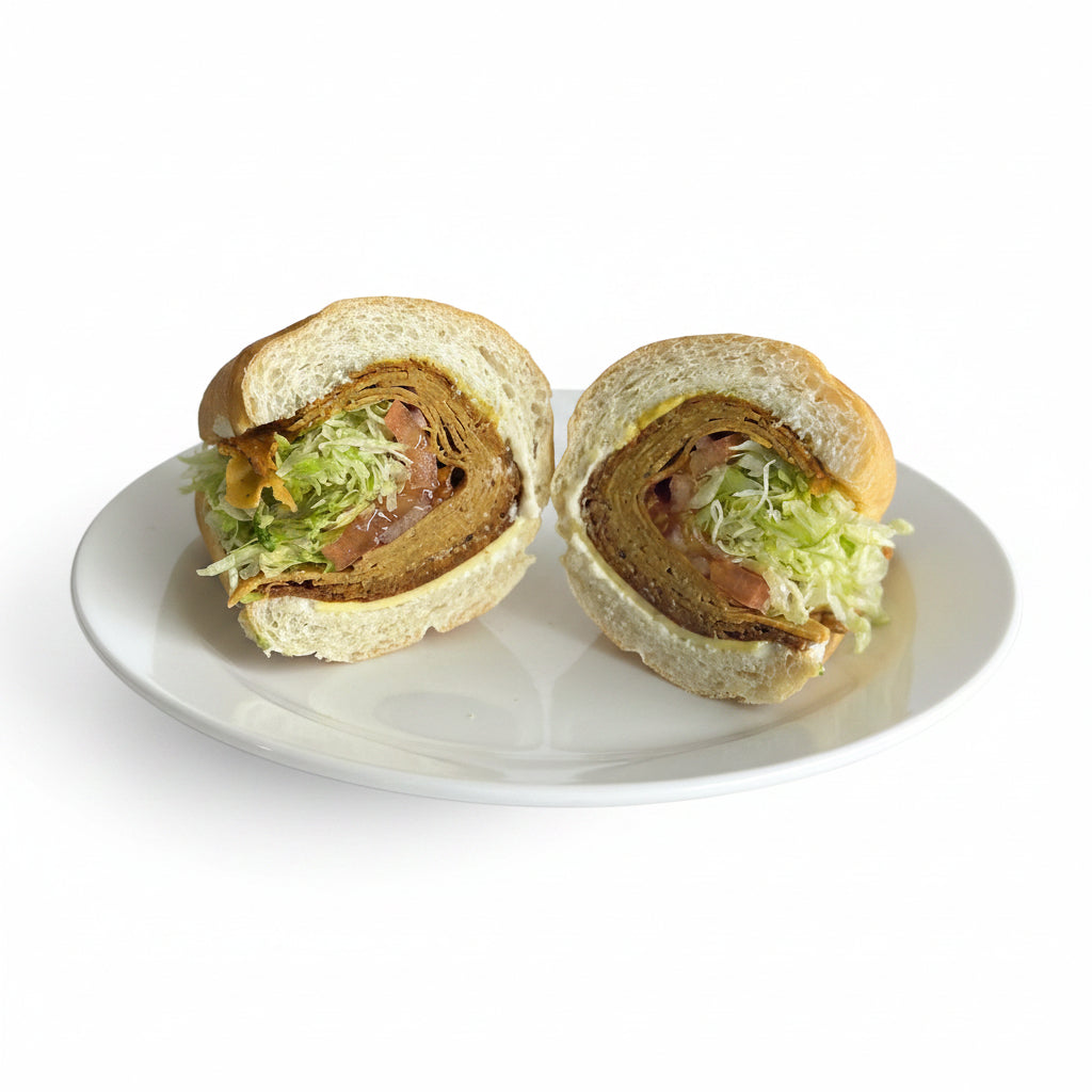 A sandwich named 'The Trio' consisting of house made corned beef, turkey and pepperoni seitan with deli mustard, horseradish mayonnaise, smoked provolone, lettuce and tomato, served on a soft grinder roll.