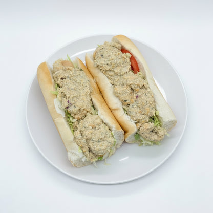 Two sandwiches with chickpea salad, nori, mayo, onion, garlic, salt, celery, lettuce, and tomato, served on a soft grinder roll, presented in a cardboard container with red and white checkered paper.